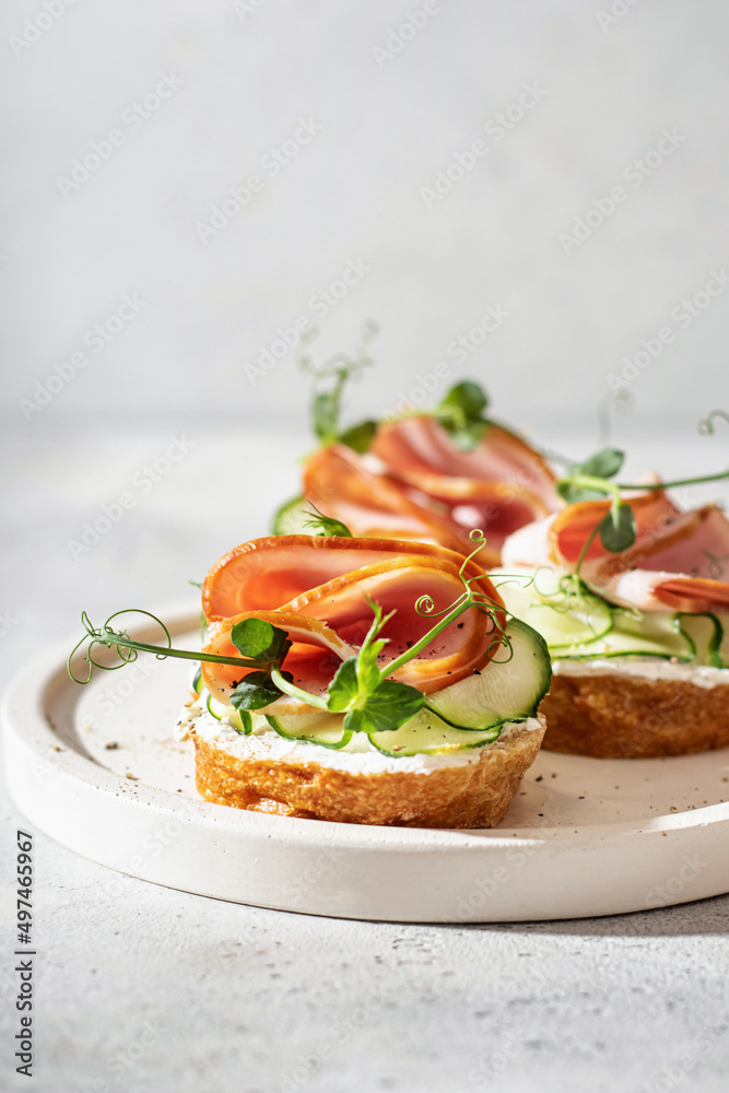 Three bruschettas with baguette, bacon or meat, cream cheese, micro-greenery, fresh cucumber and sprouts, on white plate on white textured background