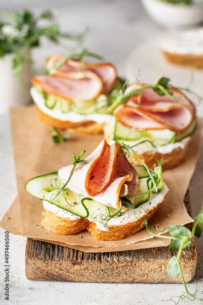Bruschettas with baguette, bacon or meat, cream cheese, micro-greenery, fresh cucumber and sprouts, in composition on wooden board on white textured background