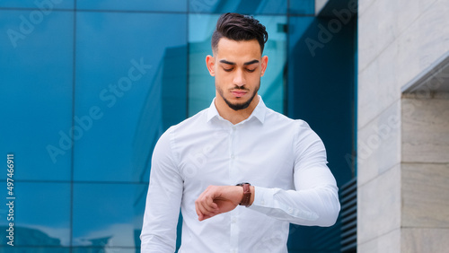 Serious upset angry hispanic indian man bearded guy disgruntled businessman boss wears formal stylish shirt stands outdoors looking at wrist watch worried about being late lateness time rush waiting photo