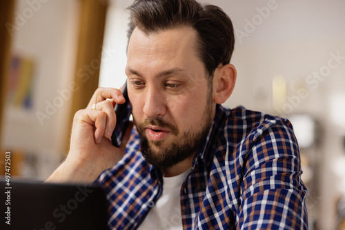 Portrait of a handsome man with a beard talking on the phone in a white blouse and blue plaid shirt, looking at a latop. Job interview. Business Phone.