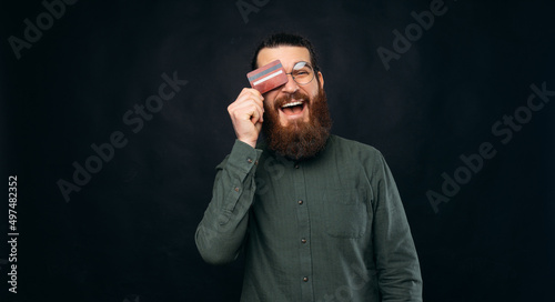 Cheerful bearded man wearing glasses is hiding eye behind a credit card. Studio shot over black background.
