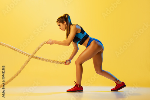 Portrait of young sportive girl training with sports ropes isolated on yellow studio background. Sport, action, fitness, youth and health concept.