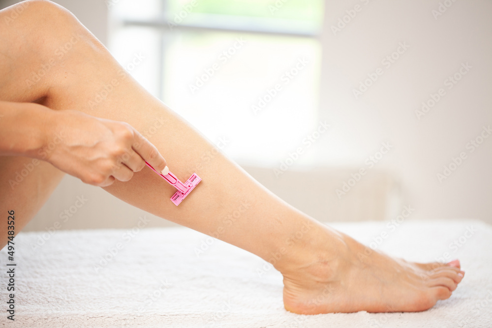 Fototapeta premium Young woman shaving legs with a razor blade in the bathroom