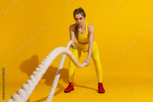 Studio shot of sportive slim girl workout with sports equipment isolated on bright yellow studio background with shadow. Beauty, sport, action, fitness, youth concept.