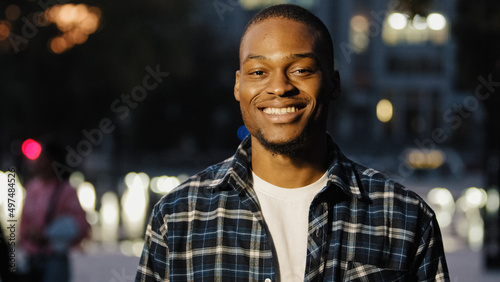 Portrait happy african american 20s millennial satisfied man guy stands in city in evening background looking at camera smiling toothy dental posing outdoors late time male face close up