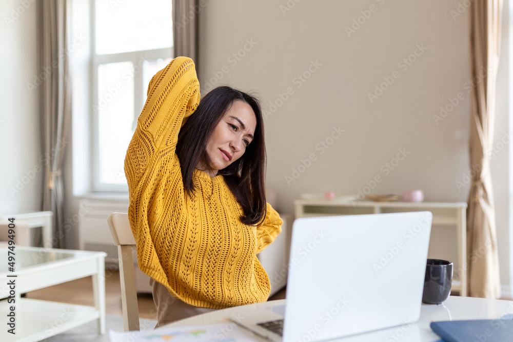Portrait of young stressed woman sitting at home office desk in front of laptop, touching aching back with pained expression, suffering from backache after working on laptop