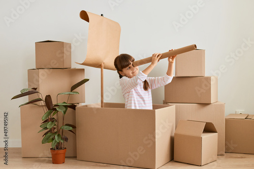 Charming dark haired little girl playing in handmade ship from cardboard box with sail, female child with spotting scope, posing surrounded with carton parcels.