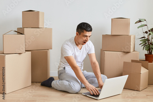 Portrait of shocked surprised man wearing casual clothing sitting on floor near carton parcels and typing on notebook keyboard, expressing fear and astonishment, forgot about important task.