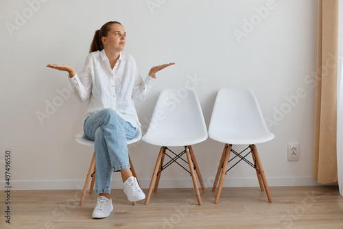 Photography Horizontal shot of helpless confused woman wearing white shirt and jeans sitting on chair in queue, spreading hands, shrugging shoulders, expressing puzzlement