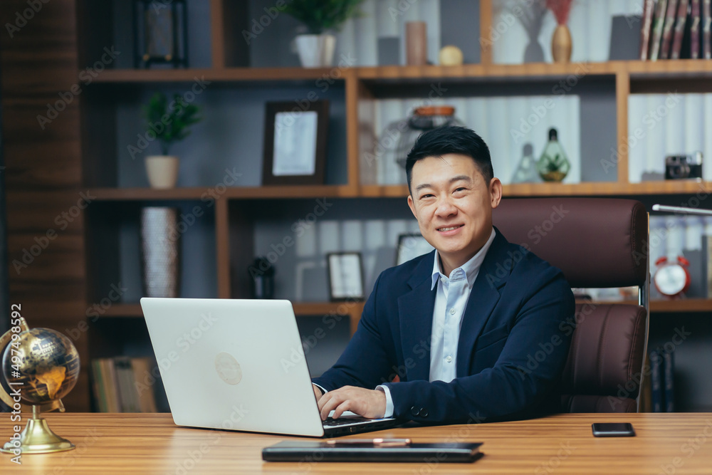 Portrait of a successful Asian businessman, man working in the office sitting at the table, looking at the camera and smiling, happy banker with laptop