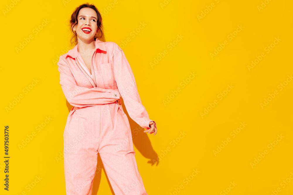 Young beautiful smiling female in trendy summer pink overalls. Sexy carefree woman posing near yellow wall in studio. Positive model having fun and going crazy. Cheerful and happy Young beautiful smiling female in trendy summer pink overalls. Sexy carefree woman posing near yellow wall in studio. Positive model having fun and going crazy. Cheerful and happy