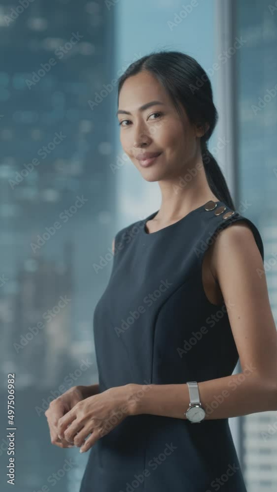 Vertical Portrait of an Asian Businesswoman in Stylish Black Dress Using Laptop Computer, Posing Next to Window in City Office. Confident Female CEO Smiling. Successful Diverse Business Manager. Vertical Portrait of an Asian Businesswoman in Stylish Black Dress Using Laptop Computer, Posing Next to Window in City Office. Confident Female CEO Smiling. Successful Diverse Business Manager.