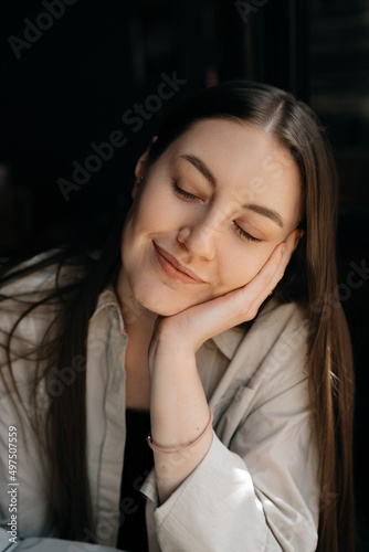 Portrait of a young European girl with long dark hair in a beige shirt with her eyes closed. She feels calm and happy