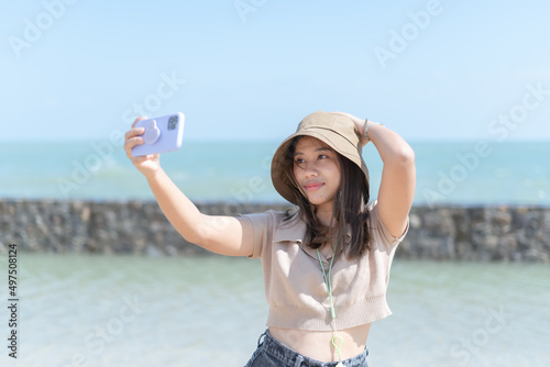 Joyful young adult Asian female tourist, traveler, woman using smartphone for selfie, taking photo at tourism, sea. Woman sharing her traveling on social media online platform. traveling concept.