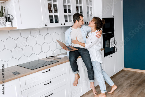 Joyful stylish husband and wife using laptop, watching funny videos or photos, smiling. Happy family couple are standing at the kitchen at home doing online shopping or chatting with friends