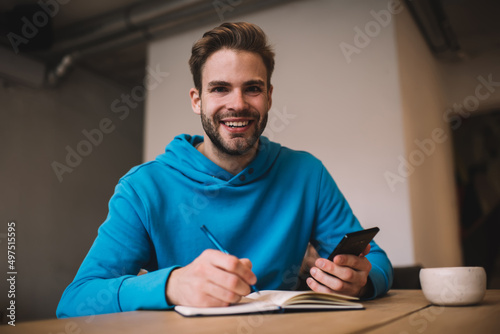Portrait of happy male student enjoying leisure time for creating content ideas using cellular gadget, cheerful hipster guy with education textbook and digital smartphone device smiling at camera