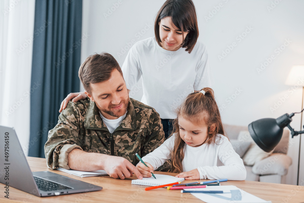 Sitting by the table. Soldier in uniform is at home with his wife and daughter