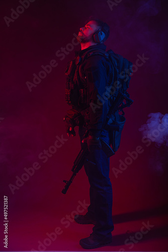 soldier in full gear with weapons. a man in headphones, body armor, with a backpack and a belt. red background. colored, blue-red light. smoke around the military. explosion, chemical attack © Ольга Новицкая