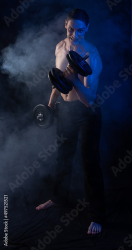 bare-chested man in colored light. showing muscles posing with dumbbells and smoke. standing or on the floor. Black background.