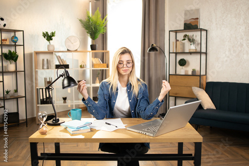 Blond woman freelancer in eyeglasses sitting at table with closed eyes and relieving stress by meditation at workplace. Concept of relaxation and harmony, no stress free relief at work