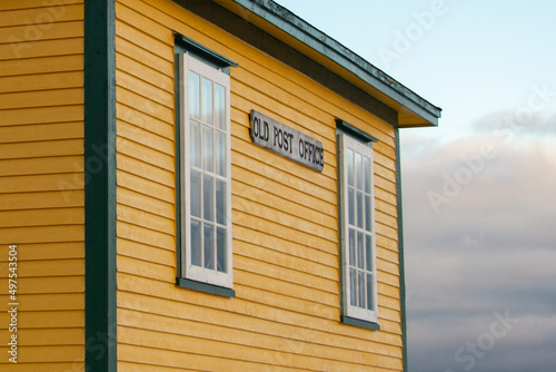 A wooden sign with black letters spelling old post office affixed to the exterior of a yellow building with green trim. There are vintage glass windows with white trim on the corner of the building. 