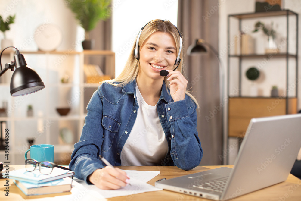 Pretty young Caucasian woman in headset making notes and smiling, while using laptop, working or studying online at bright modern room at home. Distance work and education during pandemy Pretty young Caucasian woman in headset making notes and smiling, while using laptop, working or studying online at bright modern room at home. Distance work and education during pandemy