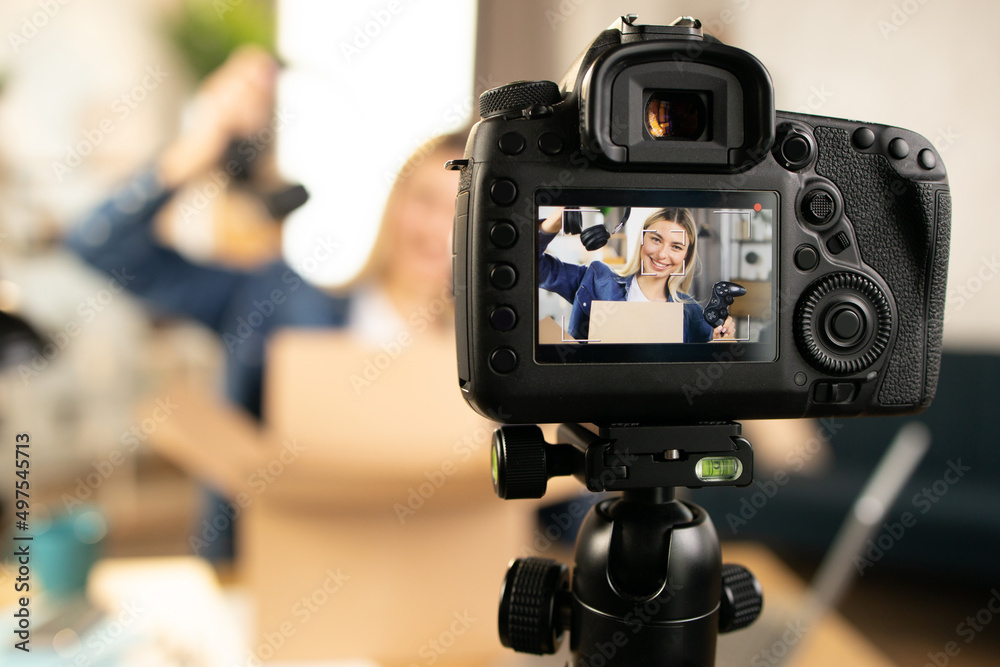 Close up of modern video camera with smiling Caucasian woman on screen. Young female influencer doing live stream while unpacking box with new headphones and joystick for games. Close up of modern video camera with smiling Caucasian woman on screen. Young female influencer doing live stream while unpacking box with new headphones and joystick for games.