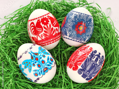 Obraz na płótnie Four Colourful Easter Eggs with floral, birds, people folk art pattern printed on stickers. Eggs in green paper grass nest. Polish traditional paper-cut from Kurpie region - Wycinanka Kurpiowska. w salonie