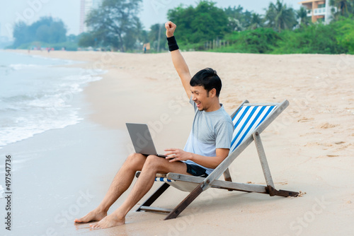 A handsome Asian man is chatting with his supervisor on a laptop, chilling in a chair by the beach, and has a happy expression on the job well done,work from home.
