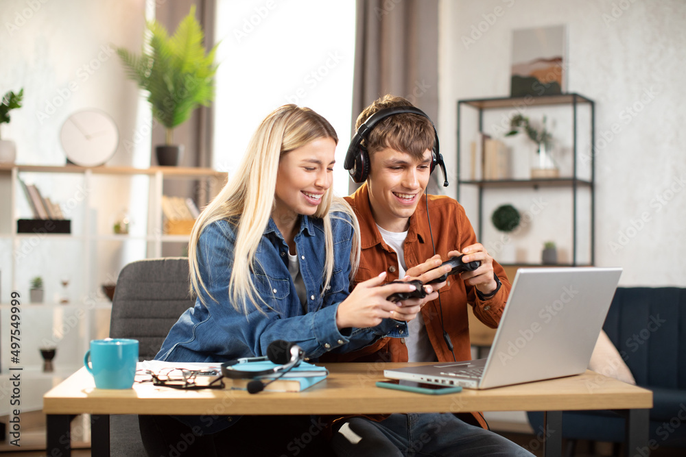 Young happy couple playing video games at home, sitting at the table with laptop and using joysticks. Happy man in headset shows new game for his lovely smiling girlfriend ar home Young happy couple playing video games at home, sitting at the table with laptop and using joysticks. Happy man in headset shows new game for his lovely smiling girlfriend ar home