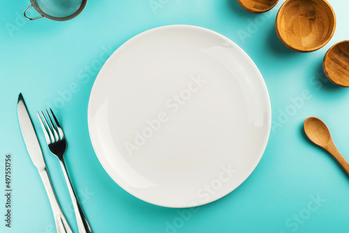 Empty white plate on the green table. Top view. Kitchen utensils beside like fork, knife, bamboo containers