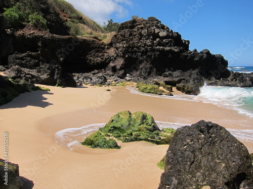 Black rocks surrounding a deserted beach. A rock with green moss at the center. View of the blue sky with clouds and waves. Nobody. Waimea, North Shore, Oahu, Hawaii. Beige, brown sand.