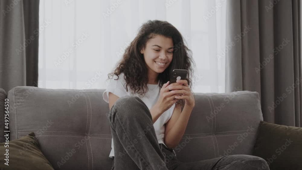 Happy millennial African American girl sitting on a couch with her phone. Smiling and texting cheerfully Happy millennial African American girl sitting on a couch with her phone. Smiling and texting cheerfully