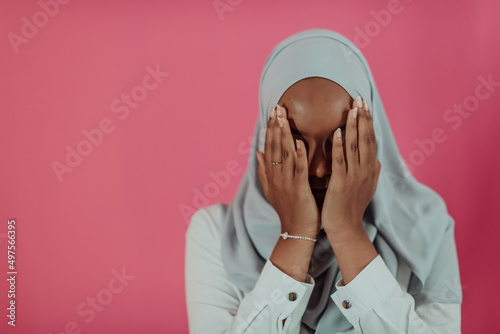 Modern African Muslim woman makes traditional prayer to God, keeps hands in praying gesture, wears traditional white clothes, has serious facial expression, isolated over plastic pink background