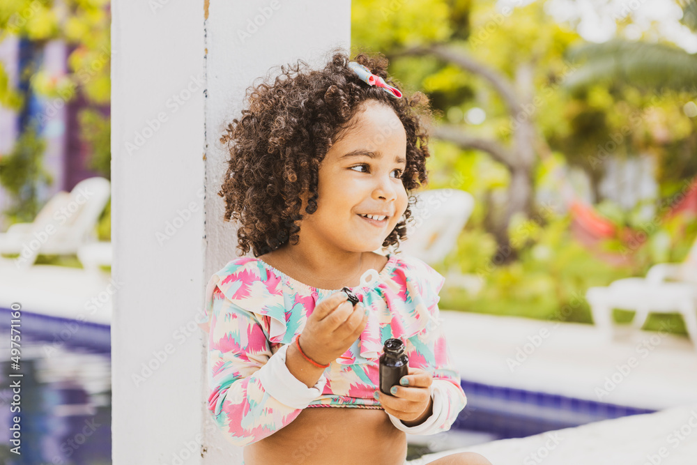© EZ PHOTOS - Portrait of smiling baby girl with afro hair style and sunglasses in a colourful swimsuit outfit blowing a bubble outdoors. Afro-American and Afro-Brazilian children summer concept with copy space. © EZ PHOTOS - Portrait of smiling baby girl with afro hair style and sunglasses in a colourful swimsuit outfit blowing a bubble outdoors. Afro-American and Afro-Brazilian children summer concept with copy space.