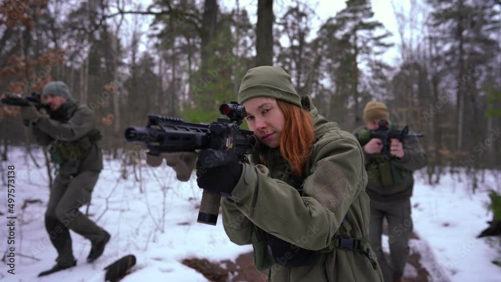 Beautiful young Caucasian woman in camouflage with strikeball gun walking in slow motion aiming as blurred troop strolling behind. Portrait of concentrated attractive female soldier training in forest Beautiful young Caucasian woman in camouflage with strikeball gun walking in slow motion aiming as blurred troop strolling behind. Portrait of concentrated attractive female soldier training in forest