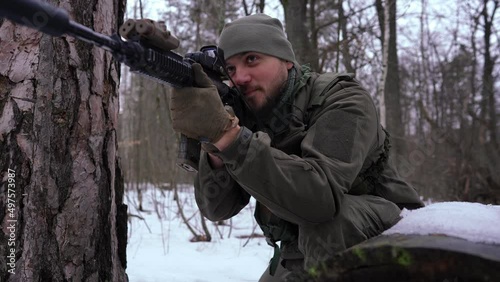 Portrait of young man with strikeball gun imitating shooting in slow motion outdoors. Handsome armed Ukrainian soldier in winter forest training. Lifestyle and defense concept photo