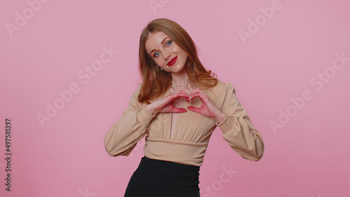 Smiling lovely businesswoman girl in beige blouse makes sign heart shape gesture demonstrates love expresses good feelings and sympathy. Young adult woman isolated alone on pink studio background © Andrii Iemelianenko