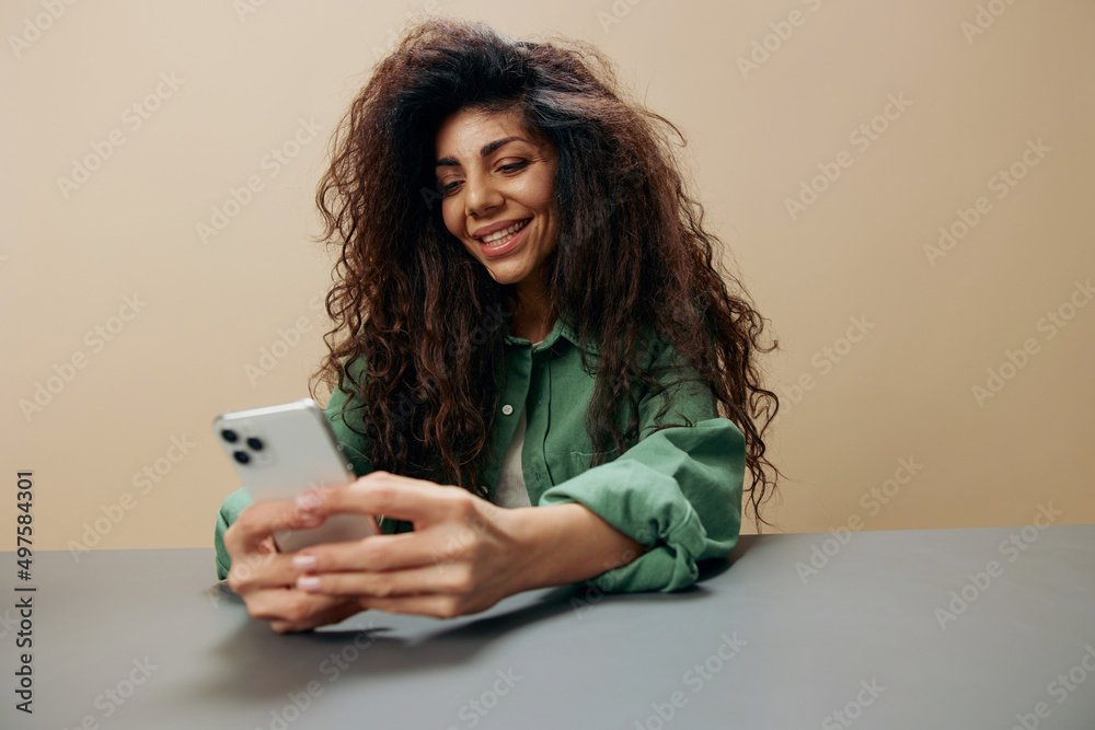 Enjoyed cheerful tanned curly Latin lady chatting with lover friends play game sit at the table with phone isolated over pastel beige background. Copy space Mockup Banner. Concept Mobile App Enjoyed cheerful tanned curly Latin lady chatting with lover friends play game sit at the table with phone isolated over pastel beige background. Copy space Mockup Banner. Concept Mobile App