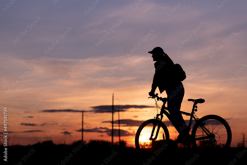 Obraz premium Bicycle rider silhouette with sunset sky in background, girl riding her bike staring at the sunny evening sky