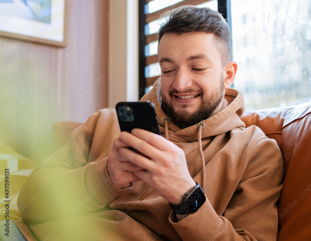 Smiling man holding phone, browsing apps, sitting on couch and spending leisure time with mobile device, chatting in social network. Smiling man holding phone, browsing apps, sitting on couch and spending leisure time with mobile device, chatting in social network.