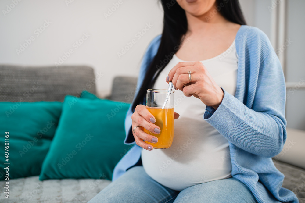 A pregnant black-haired caucasian in advanced pregnancy sits on the sofa in the sitting room and holds a glass of fruity juice. She mixes the juice with a teaspoon. Orange liquor is in the glass. A pregnant black-haired caucasian in advanced pregnancy sits on the sofa in the sitting room and holds a glass of fruity juice. She mixes the juice with a teaspoon. Orange liquor is in the glass.