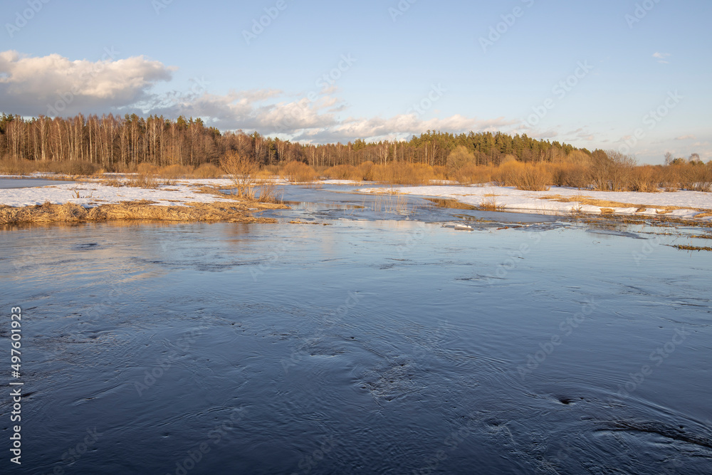 March sunny evening by the river. Blue sky over the horizon. A picturesque landscape, early spring, a river with snow-covered banks, dry grass and bushes. The first thaws, the snow is melting