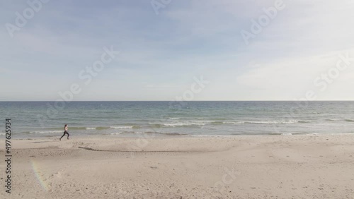 Young healthy woman jogging on tropical beach by the ocean horizon. Jogging from left to right sunny warm afternoon in slow motion. Blue sky green water bright white sand sun flare. Wide static shot. photo