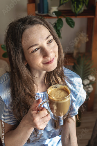Young woman is sitting by the window and drinking coffee enjoying a sunny morning. Concept of a pleasant pastime, good mood, harmony and happiness in small things