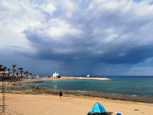 The sunny sandy coast of the Mediterranean Sea, the beach in the bay, the temple of St. Nicholas the Wonderworker, a dramatic sky and rain over the sea, a ship in the sea.