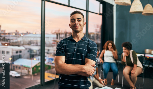 He has big dreams for their small business. Portrait of a cheerful young man standing with his arms folded with his work colleagues grouped together in the background.