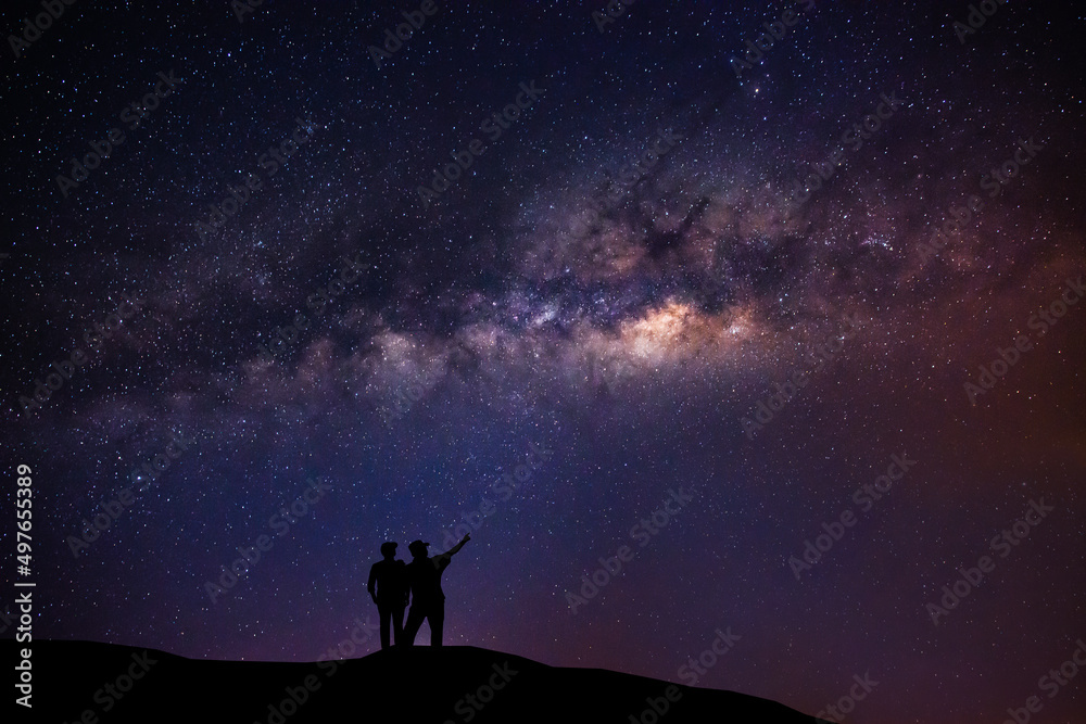A couple standing on top of a mountain next to the Milky Way galaxy pointing at a bright star.