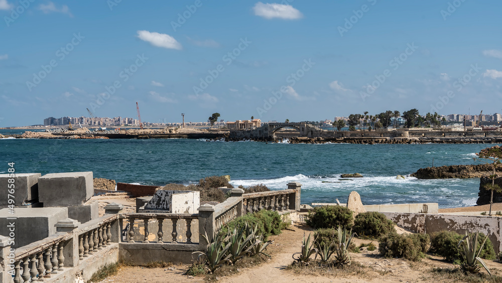 The embankment in the park of Alexandria. An old balustrade is visible, plants on dry soil. The waves of the turquoise Mediterranean Sea are foaming. City buildings in the distance. Egypt