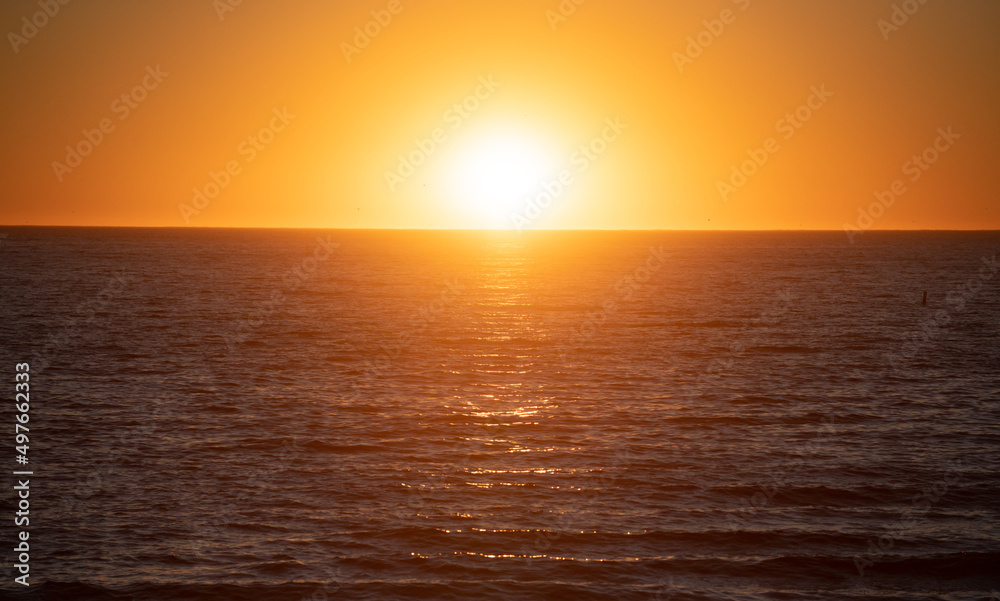 Fototapeta premium Sunset with large yellow sun under the sea surface. Calm ocean with sunset sky and sun through the clouds over. Calm ocean and sky background.